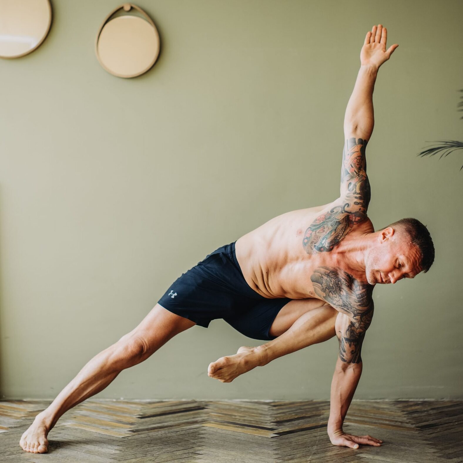 A shirtless man with tattoos performing a yoga pose indoors, showcasing flexibility and strength.